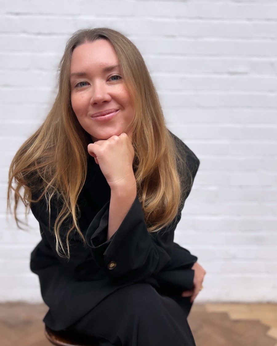 Dani Forword, seated on a wooden stool in a black suit against a white brick wall, chin resting on her hand